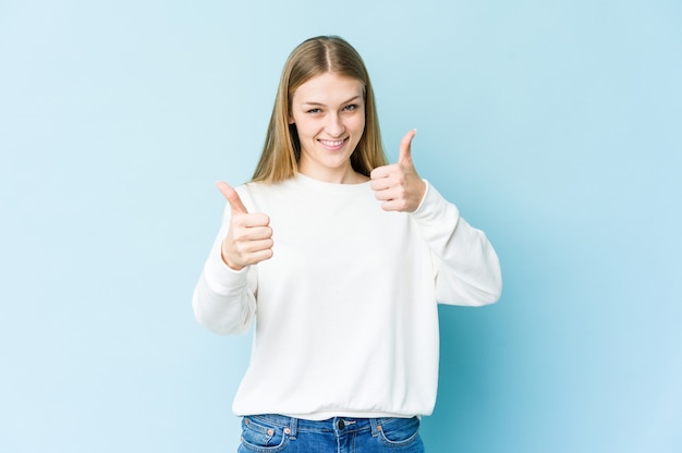 Joven rubia aislada sobre fondo azul levantando ambos pulgares, sonriente y confiada.