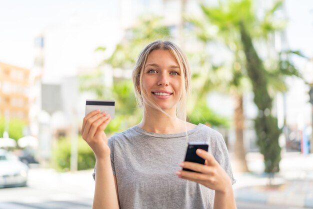 Joven rubia al aire libre comprando con el móvil con tarjeta de crédito