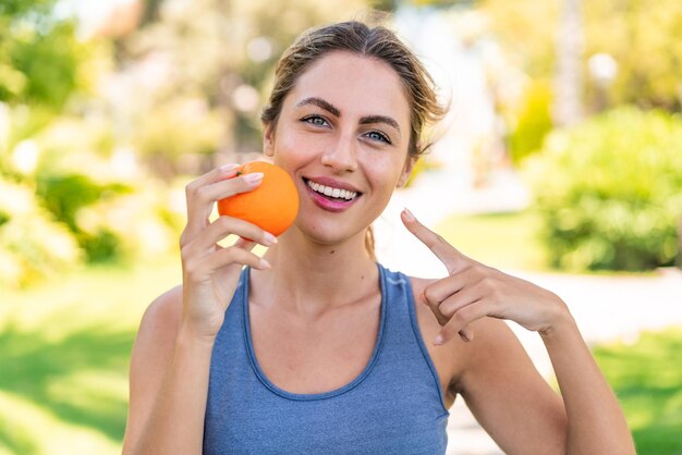 Joven rubia al aire libre sosteniendo una naranja con expresión feliz