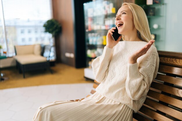 Una joven rubia alegre y emocionada hablando por teléfono móvil sonriendo discutiendo las últimas noticias disfrutando de una agradable conversación sentada en un banco de madera