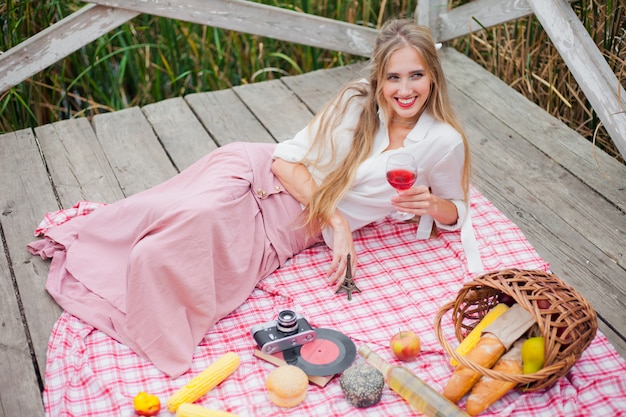 Joven rubia alegre en ropa vintage beber vino tinto mientras se encuentra en mantel de picnic en el muelle de madera