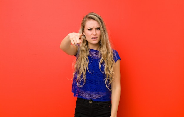 Joven rubia apuntando a la cámara con una expresión agresiva enojada que parece un jefe furioso y loco contra la pared roja