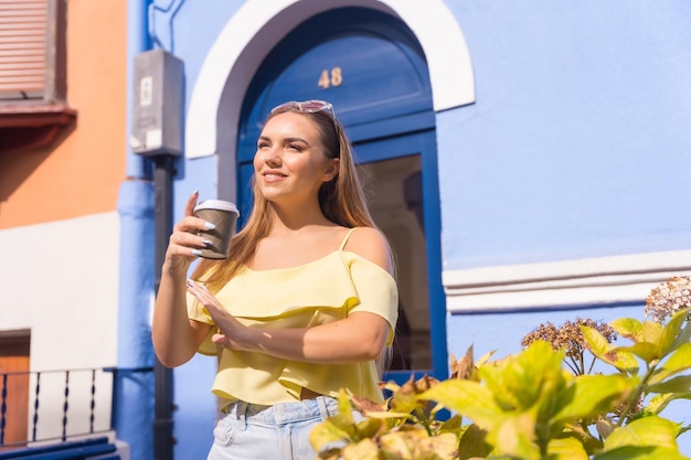 Joven rubia en un barrio de casas con fachadas coloridas con un café en la puerta de su casa