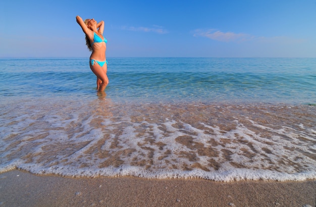 Joven rubia en bikini azul de pie en agua de mar quieta y disfrutando del sol