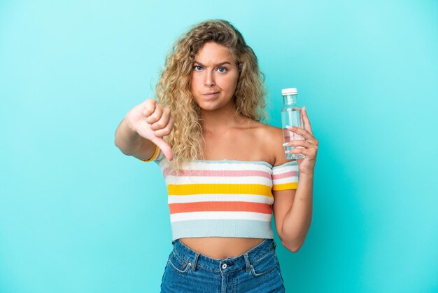 Joven rubia con una botella de agua aislada de fondo azul mostrando el pulgar hacia abajo con expresión negativa