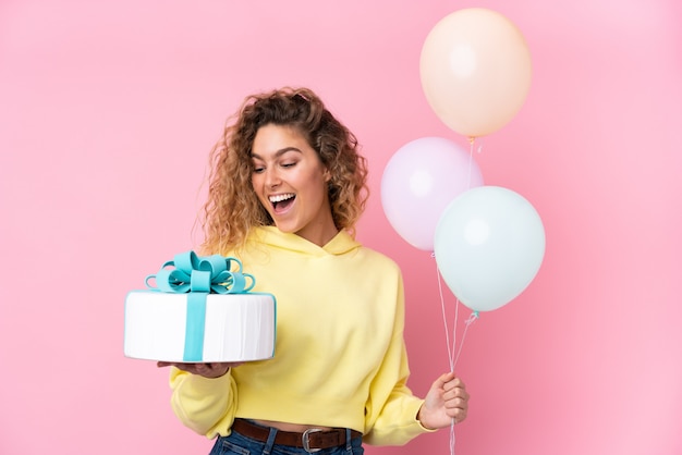 Joven rubia con cabello rizado atrapando muchos globos y sosteniendo un gran pastel en la pared rosa