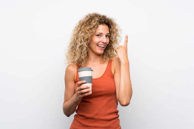 Joven rubia con cabello rizado sosteniendo un café para llevar con la intención de darse cuenta de la solución mientras levanta un dedo