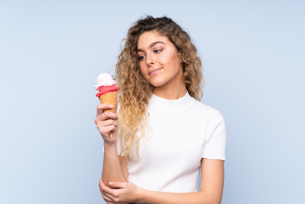 Joven rubia con cabello rizado sosteniendo un helado de cucurucho en la pared azul con expresión triste