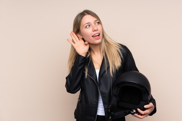 Joven rubia con un casco de moto sobre pared aislada escuchando algo