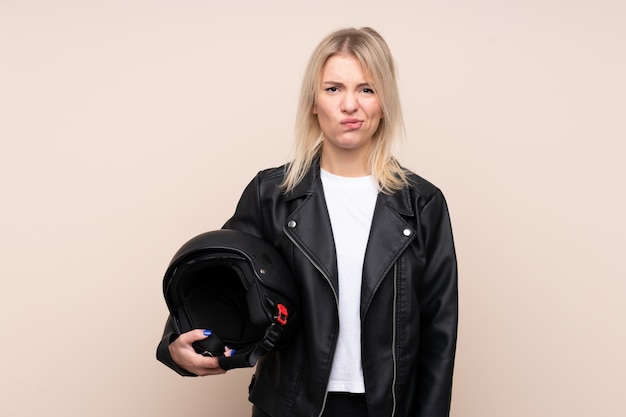 Joven rubia con un casco de moto sobre pared aislada con expresión triste