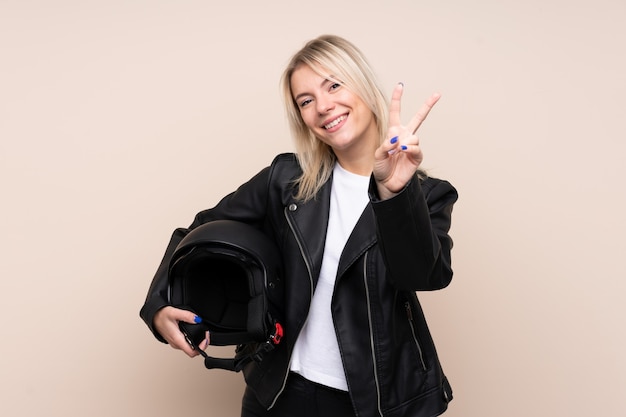 Joven rubia con un casco de moto sobre pared aislada sonriendo y mostrando el signo de la victoria