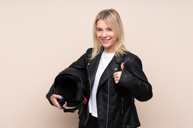 Joven rubia con un casco de motocicleta sobre una pared aislada con los pulgares arriba porque algo bueno ha sucedido