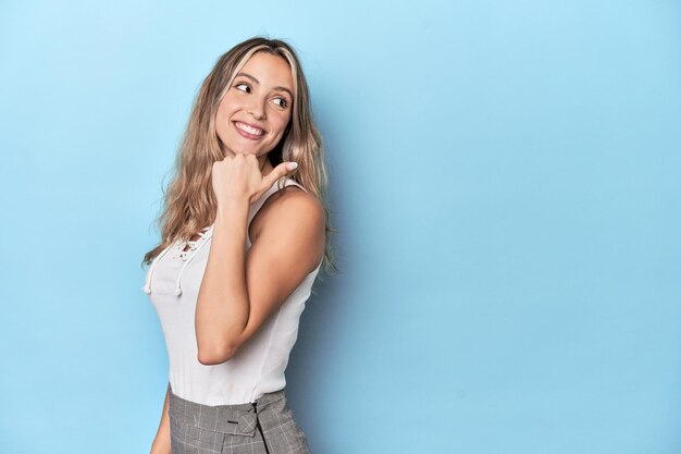 Una joven rubia caucásica en un estudio azul señala con el dedo pulgar lejos riendo y despreocupada