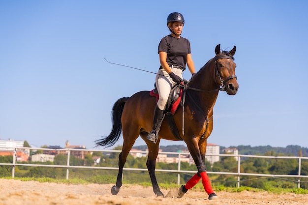 Joven rubia caucásica montando a caballo con un caballo marrón, vestida de jinete negro con gorro de seguridad