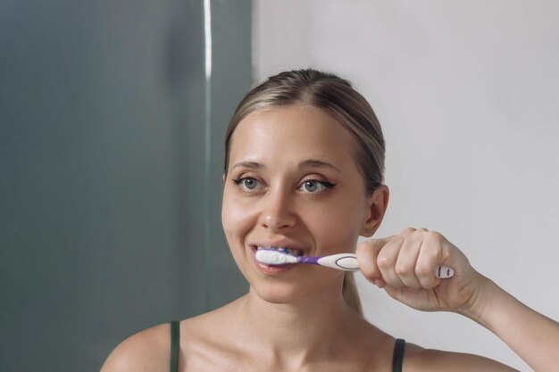 Foto una joven rubia cepillándose los dientes con un cepillo de dientes mirando en el espejo del baño