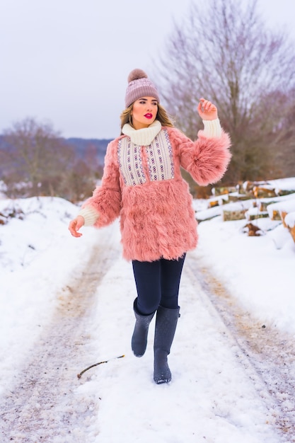 Joven rubia con chaqueta de piel rosa, botas de invierno y un sombrero morado en la nieve. Caminando por un camino lleno de nieve, estilo de vida