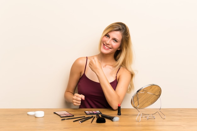 Joven rubia con cosméticos en una mesa celebrando una victoria