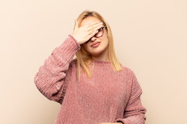 Joven rubia cubriendo los ojos con una mano sintiéndose asustada o ansiosa, preguntándose o esperando ciegamente una sorpresa