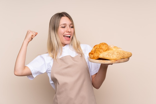 Joven rubia con delantal. Panadero hembra sosteniendo una mesa con varios panes celebrando una victoria