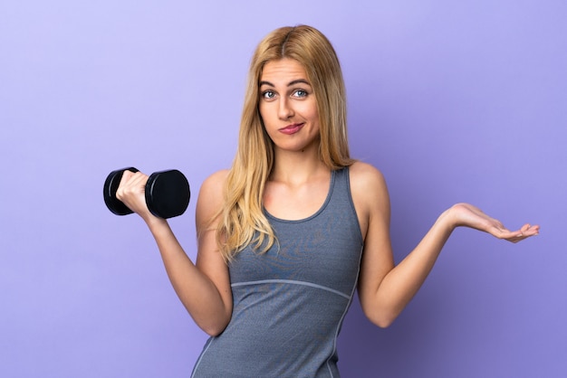 Joven rubia deporte mujer haciendo pesas sobre la pared púrpura haciendo dudas gesto mientras levanta los hombros