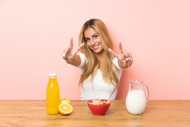 Joven rubia desayunando leche sonriendo y mostrando el signo de la victoria