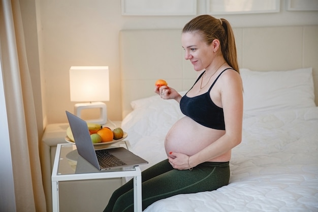 Una joven rubia embarazada está sentada en la cama y comiendo fruta mientras mira su computadora portátil. Foto de alta calidad