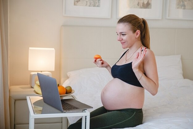 Una joven rubia embarazada está sentada en la cama y comiendo fruta mientras mira su computadora portátil