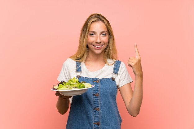 Joven rubia con ensalada sobre pared rosa aislado apuntando hacia una gran idea