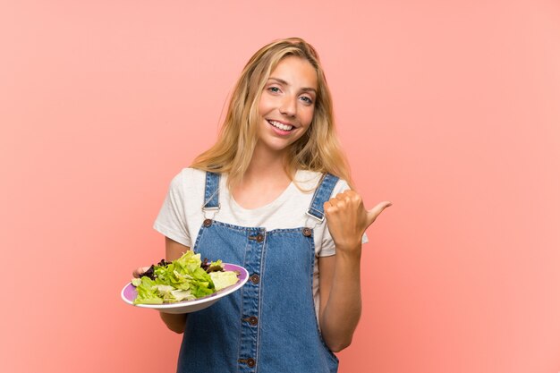 Joven rubia con ensalada sobre pared rosa aislado apuntando hacia un lado para presentar un producto