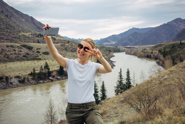 Joven rubia con gafas de sol y camiseta blanca toma un selfie en su teléfono inteligente en el fondo del río y las montañas rocosas. Feliz turista sonríe a la cámara del teléfono móvil.