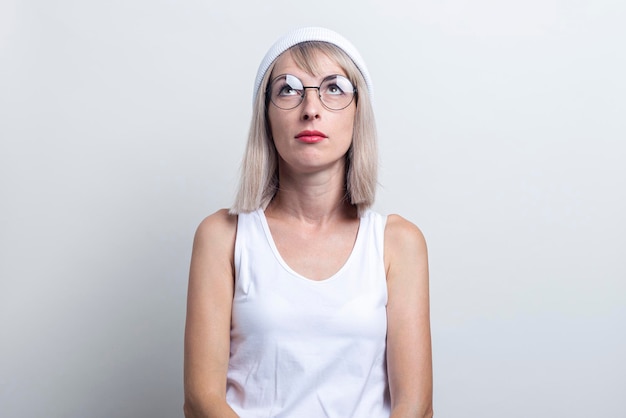 Una joven rubia con gafas y un sombrero blanco mira hacia arriba sobre un fondo claro