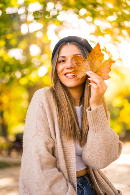 Foto joven rubia con un gorro de lana en otoño con una hoja en la cara al atardecer sonriendo