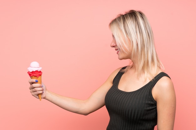 Joven rubia con un helado de cucurucho sobre pared rosa aislado con expresión feliz