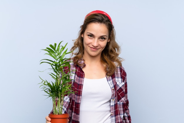 Joven rubia jardinero chica sosteniendo una planta sobre pared azul aislada