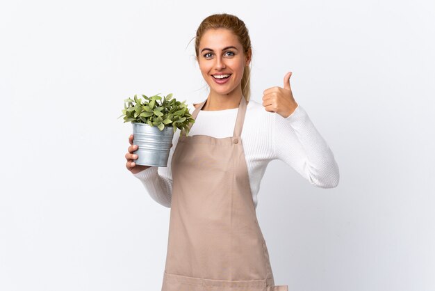 Joven rubia jardinero mujer niña sosteniendo una planta sobre blanco aislado dando un gesto de pulgares arriba