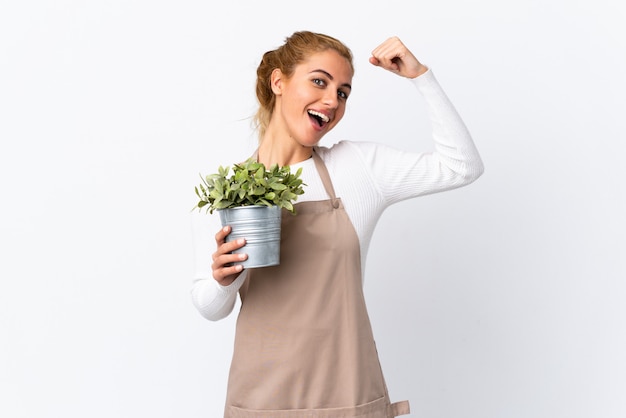 Joven rubia jardinero mujer niña sosteniendo una planta sobre blanco aislado haciendo un gesto fuerte