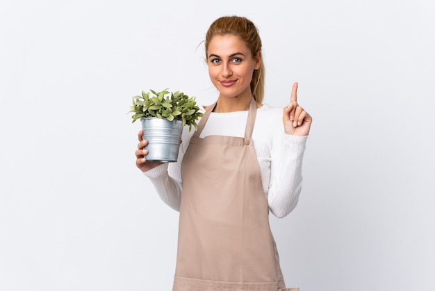 Joven rubia jardinero mujer niña sosteniendo una planta sobre blanco aislado señalando con el dedo índice una gran idea