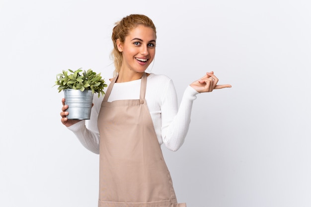 Joven rubia jardinero mujer niña sosteniendo una planta sobre pared blanca aislada sorprendido y apuntando con el dedo al lado