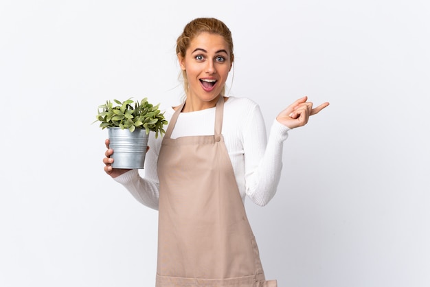 Joven rubia jardinero mujer niña sosteniendo una planta sobre pared blanca apuntando el dedo hacia un lado