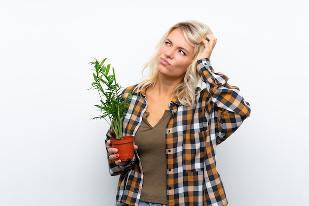 Joven rubia jardinero mujer sosteniendo una planta sobre pared blanca aislada con dudas y con expresión de la cara confusa