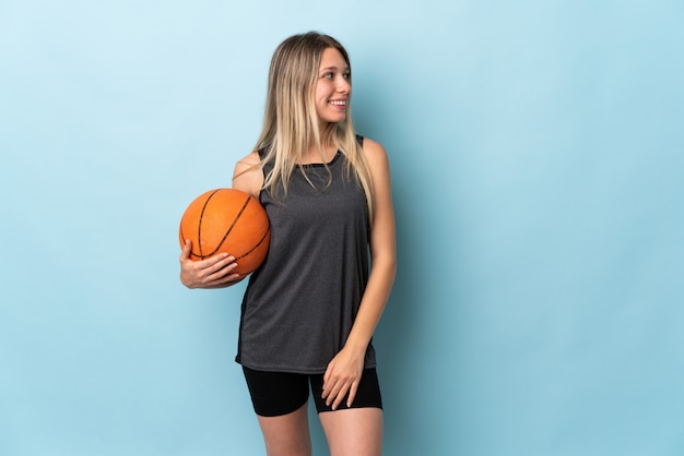 Joven rubia jugando baloncesto aislado en la pared azul mirando hacia el lado