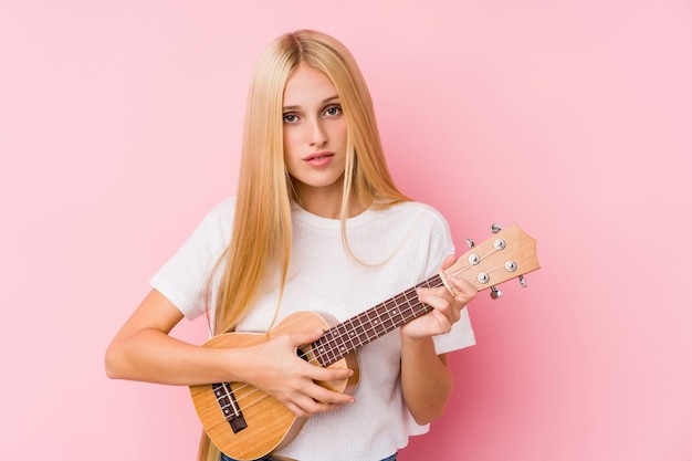 Joven rubia jugando ukelele aislado en una pared.