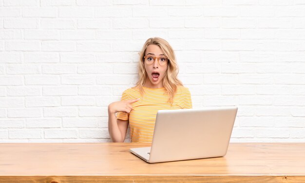 joven rubia en laptop mirando sorprendido y sorprendido con la boca abierta apuntando a sí mismo