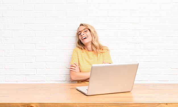 joven rubia en laptop riendo alegremente con los brazos cruzados