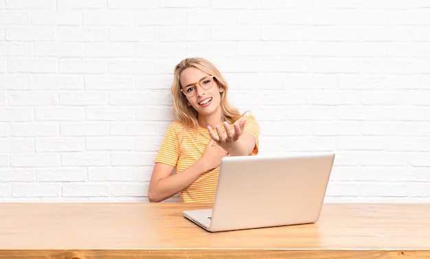 joven rubia en laptop sonriendo con una mano al lado del corazón y la otra estirada al frente