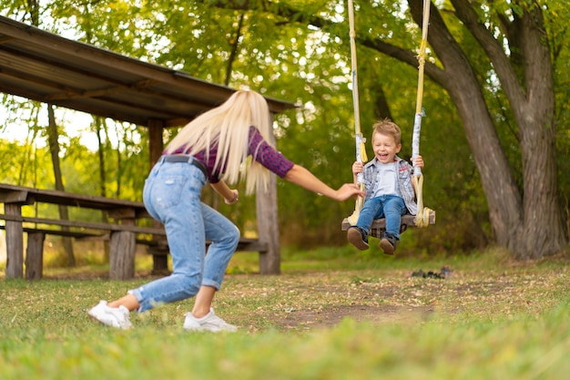 Joven rubia mamá sacude a su pequeño hijo en un columpio en un parque verde. Infancia feliz.