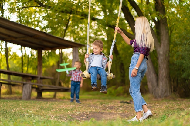 Joven rubia mamá sacude a su pequeño hijo en un columpio en un parque verde. Infancia feliz.