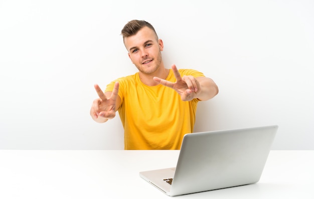 Joven rubia en una mesa con un portátil sonriendo y mostrando el signo de la victoria