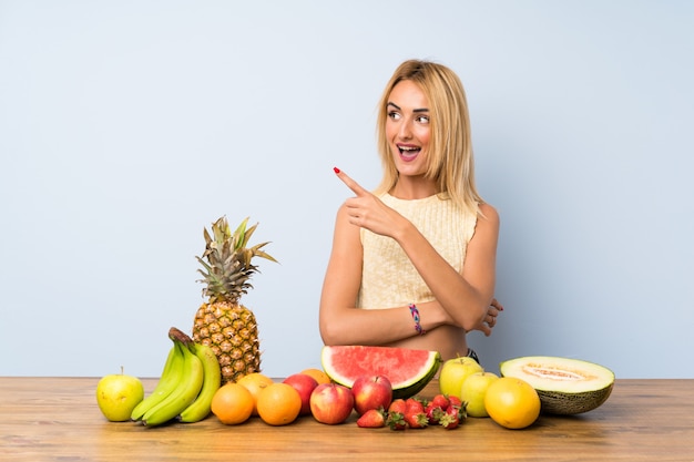 Joven rubia con muchas frutas apuntando con el dedo al lado