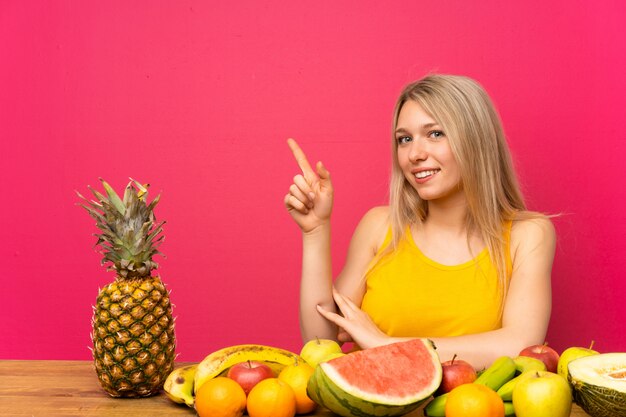 Joven rubia con muchas frutas apuntando con el dedo al lado